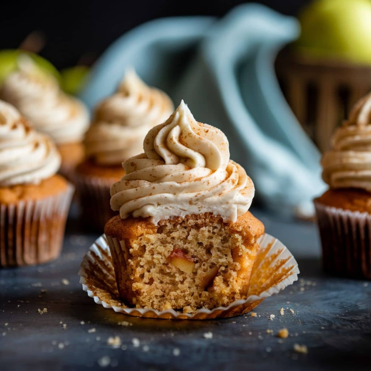 Homemade Apple Cinnamon Cupcake with cinnamon cream cheese frosting, unwrapped and placed on a dark rustic surface, with crumbs scattered around and additional cupcakes in the blurred background.