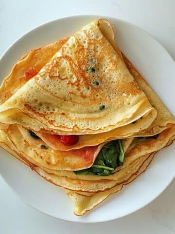 Stack of gluten free crepes folded and rolled with sweet and savory fillings on a white counter.