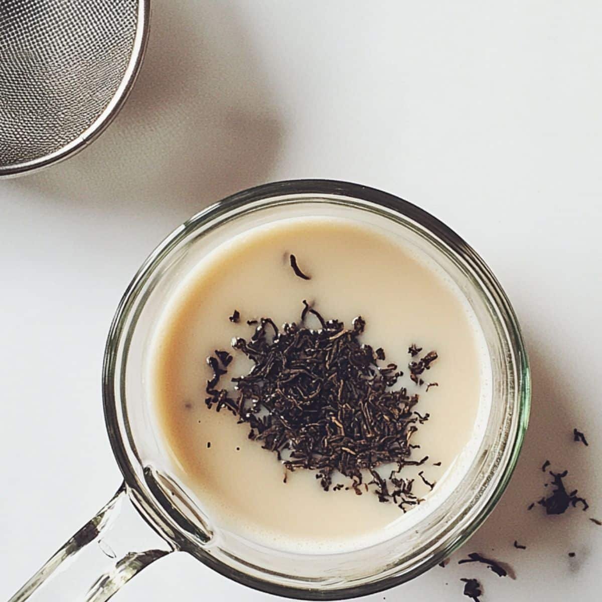 Loose Earl Grey tea steeping in warm milk with strainer beside it. Overhead shot on white kitchen counter.