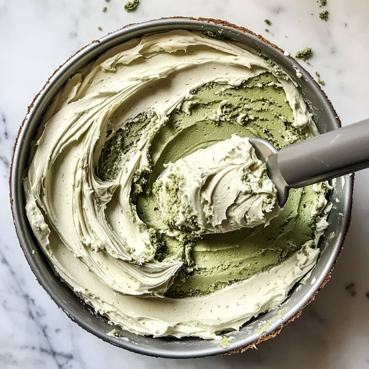 Frosting being spread on top of a vibrant green matcha cake. Thick cream swirls and crumbs visible in a real kitchen.