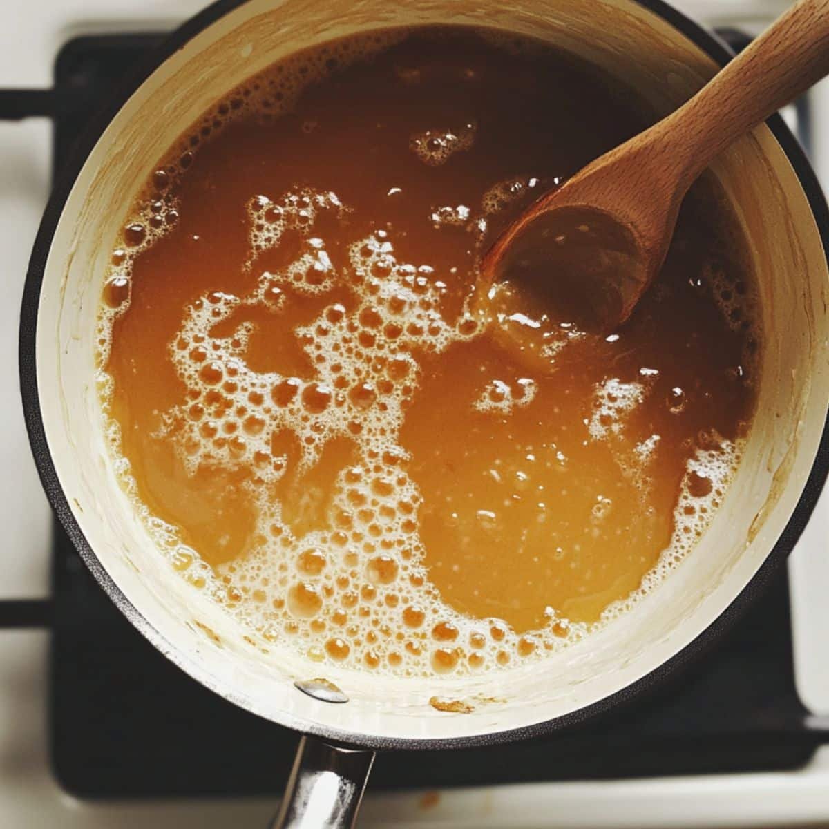 Homemade caramel sauce simmering in a saucepan, mid-cook with wooden spoon nearby on a white counter.