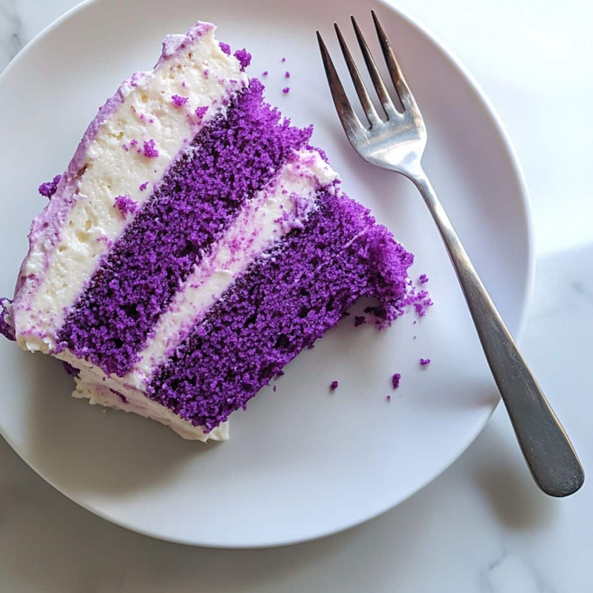 A slice of ube cake with uneven cream cheese frosting on white plate. Overhead shot with crumbs and a fork beside it.