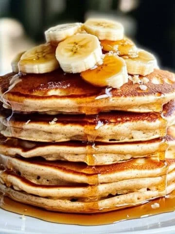 Fluffy almond flour banana pancakes stacked high with banana slices and syrup on a white plate.