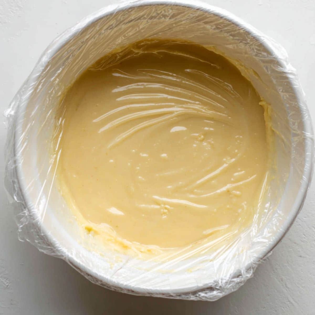 Bowl of warm pastry cream with plastic wrap on top cooling on white counter	