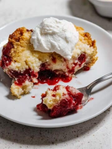 Top-down view of a rustic strawberry-cheesecake dump cake with a plated serving and whipped cream.