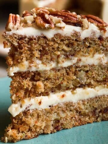 A thick, rustic slice of homemade hummingbird cake recipe with cream cheese frosting and pecans on top, on a white plate