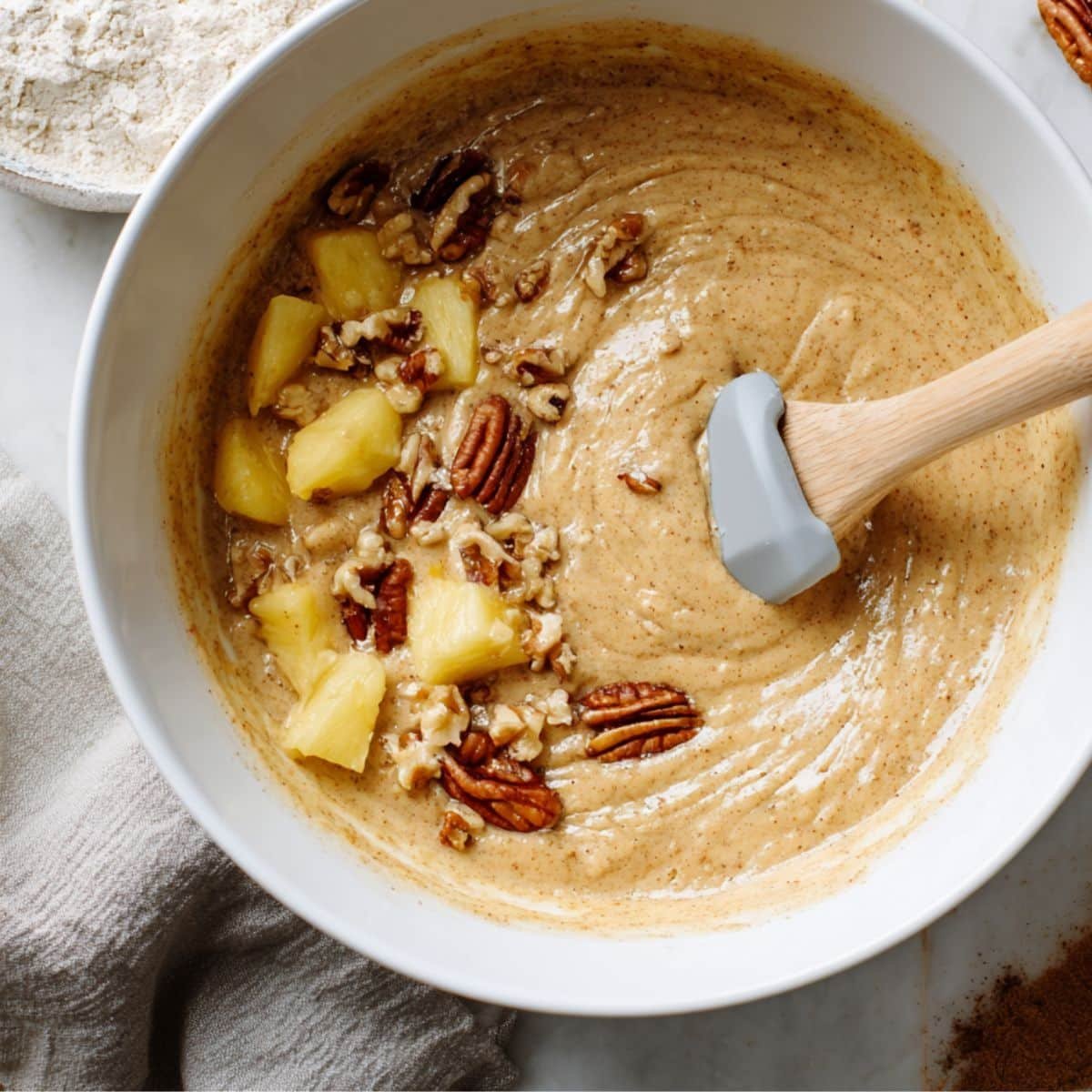 Mixing bowl filled with hummingbird cake batter being stirred with a spatula on a white counter	