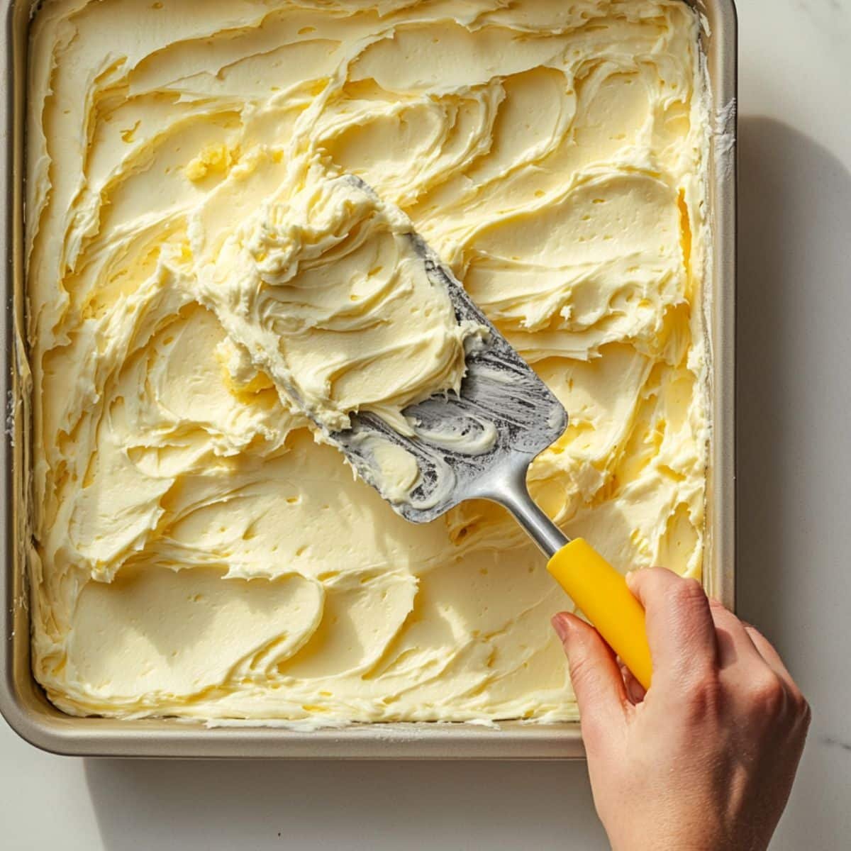 Frosting being spread on cooled cake in a 9x13 pan using spatula	