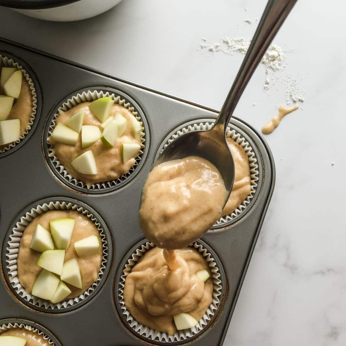Scooping chunky apple muffin batter into muffin liners on a white kitchen counter	