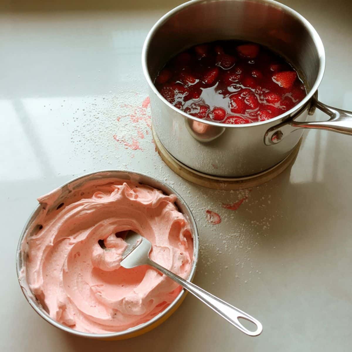 Strawberry filling in pan and pink buttercream in bowl on counter
