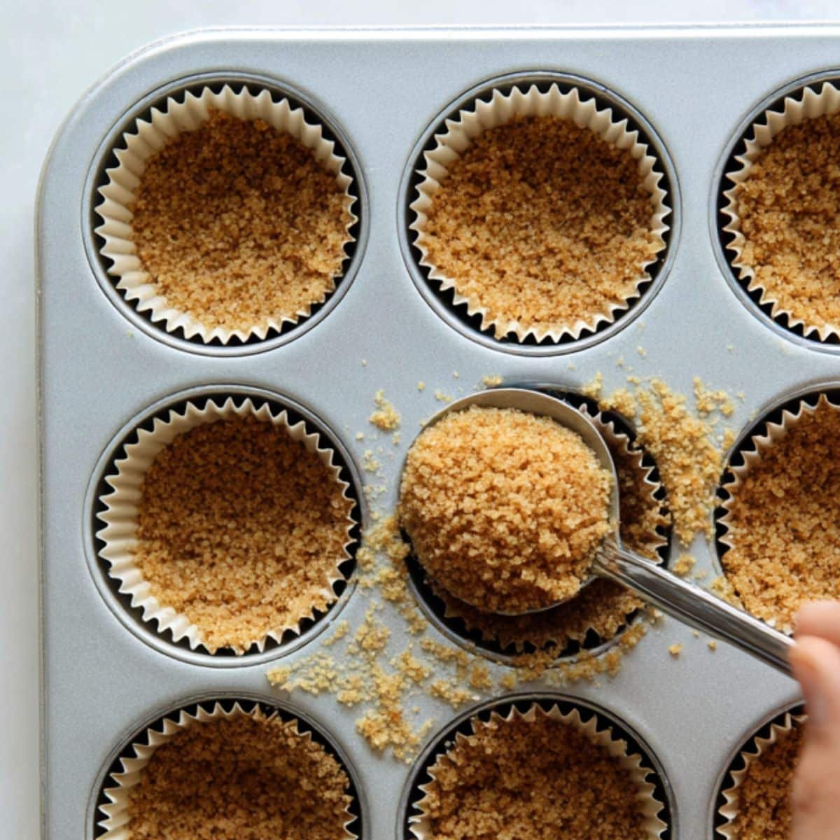 Pressing graham cracker crust into mini muffin tin on white kitchen counter	