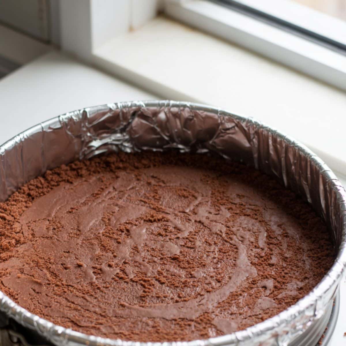 Pressing chocolate graham cracker crust mixture into a springform pan on a white counter	