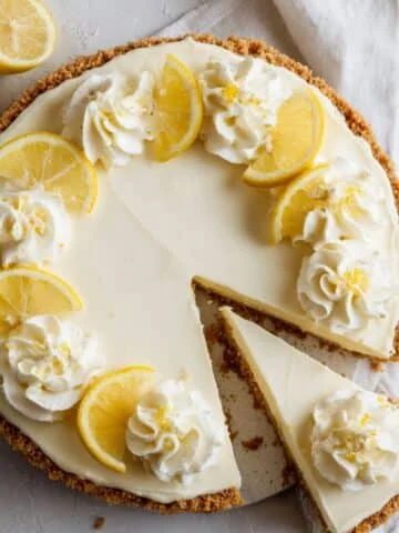 Homemade no bake lemon cheesecake with graham cracker crust, topped with whipped cream and lemon slices, on a white kitchen table.