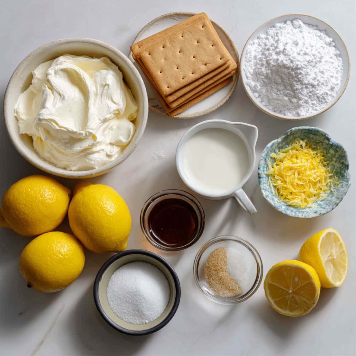 Ingredients for homemade no bake lemon cheesecake displayed on a white kitchen table: cream cheese, lemons, powdered sugar, heavy cream, vanilla extract, graham crackers, butter, sugar, salt, and lemon zest.