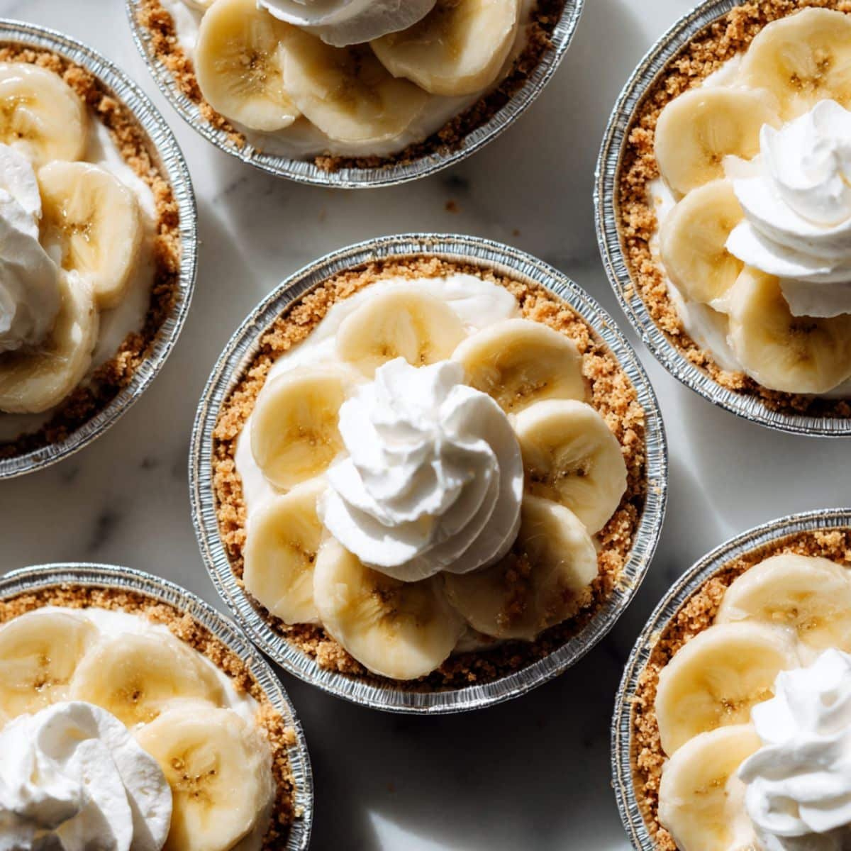 Assembling mini banana cream pies in foil tins with bananas, pudding, and whipped cream	