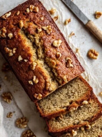 Freshly baked banana nut bread loaf with slices, topped with chopped walnuts, on a white kitchen counter.