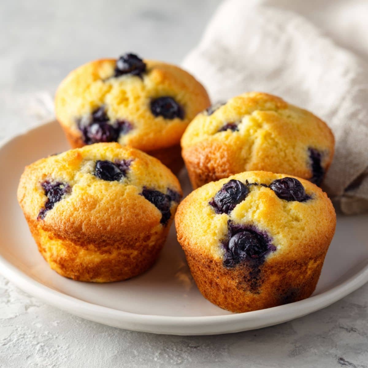 Freshly baked blueberry cornbread muffins on a white kitchen counter, golden tops with blueberries, homemade cozy look