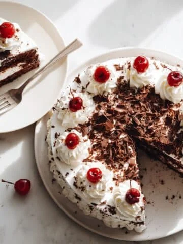 Homemade Black Forest Cake with whipped cream, cherries, and chocolate shavings, with one slice served on a plate.