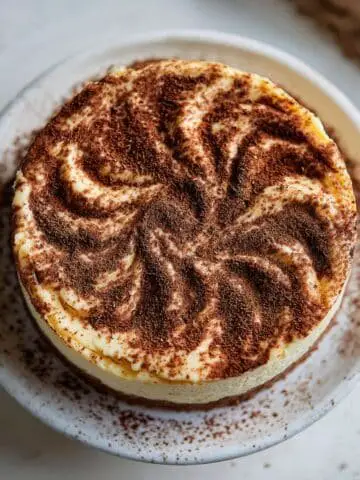 Homemade tiramisu cheesecake with cocoa dusting and cream swirls, photographed on a white kitchen table from above