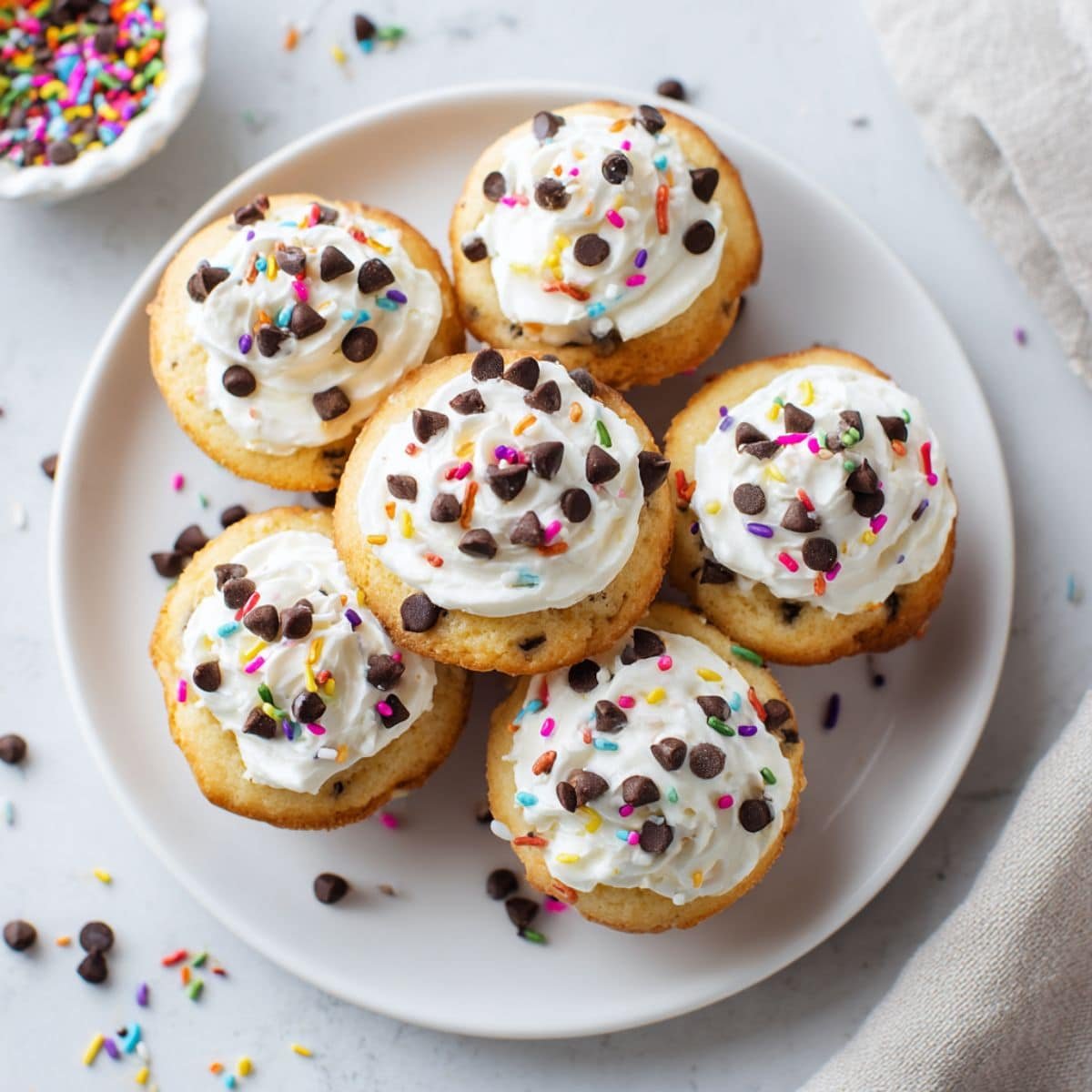 Top-down photo of homemade frosted mini cakes with sprinkles and chocolate chips on a white kitchen counter