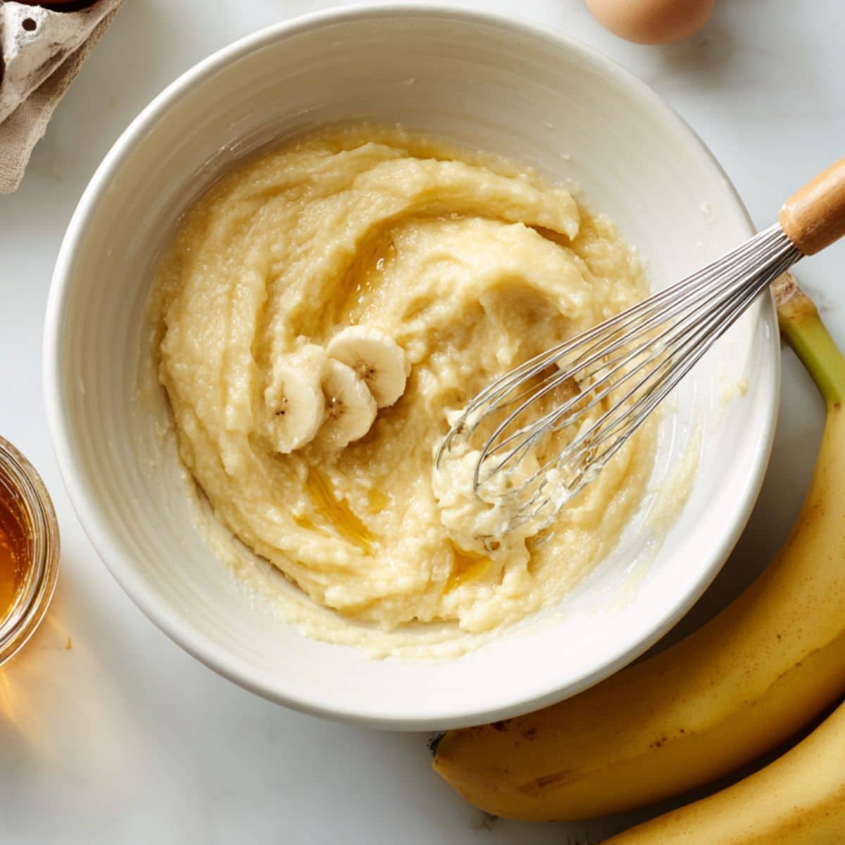 Mashed bananas in a mixing bowl with whisk and eggs nearby.	