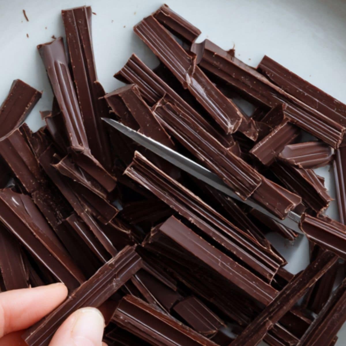 Cutting dark chocolate batons into small pieces on a white kitchen counter.