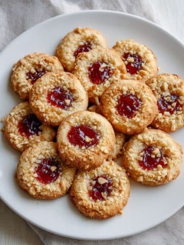 A plate of homemade Thumbprint Cookie Recipe filled with raspberry jam, rolled in chopped nuts, on a white kitchen table.
