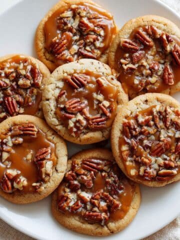 Alt text: A plate of turtle cookies with caramel centers, pecans, and chocolate drizzle on a white table.