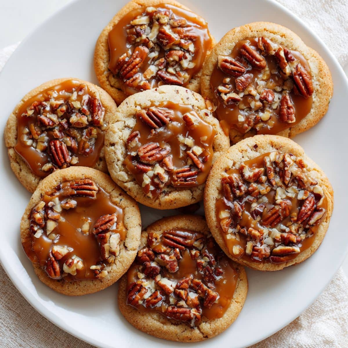 Alt text: A plate of turtle cookies with caramel centers, pecans, and chocolate drizzle on a white table.