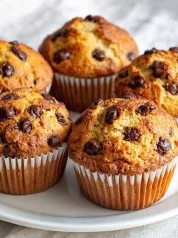 Featured image of freshly baked homemade banana chocolate chip muffins in a muffin tin on a white kitchen table, shot from above.