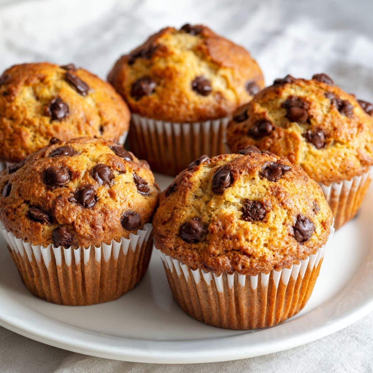 Featured image of freshly baked homemade banana chocolate chip muffins in a muffin tin on a white kitchen table, shot from above.