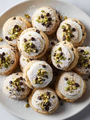 Freshly baked cannoli cookies with ricotta filling, pistachios, and powdered sugar on a white kitchen table.