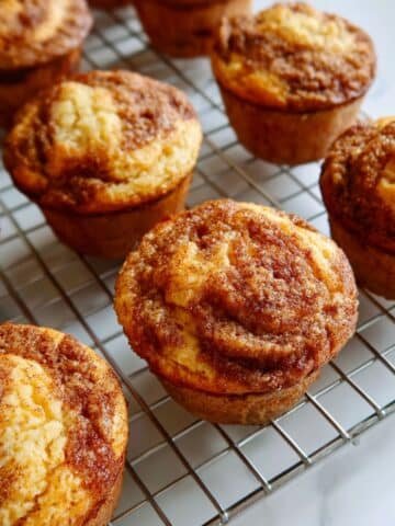 Freshly baked homemade cinnamon muffins with golden tops and cinnamon swirl, cooling on a wire rack on a white kitchen table.