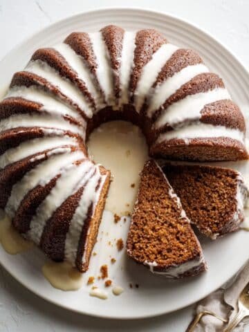 Top-down homemade gingerbread bundt cake with uneven cream-cheese glaze and one slice on a white plate, on a white kitchen table.