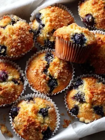 photo of a dozen blueberry Kodiak Cake Muffins on a white table with crumbs and one muffin broken open.