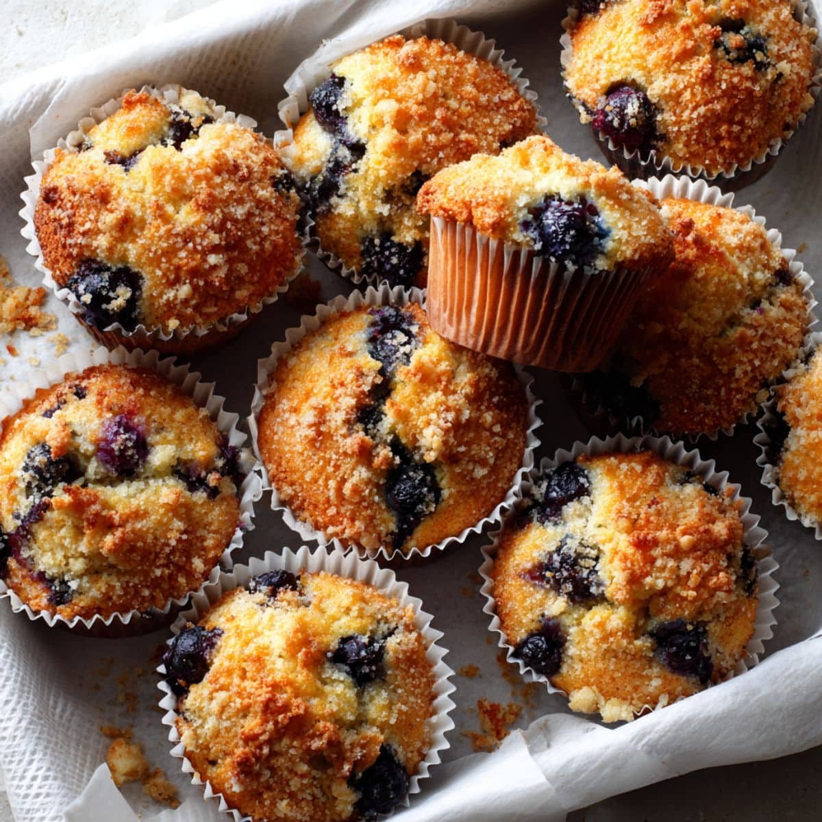 photo of a dozen blueberry Kodiak Cake Muffins on a white table with crumbs and one muffin broken open.