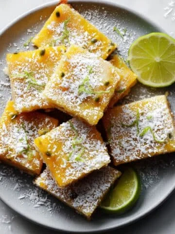 Homemade passionfruit bars cut into squares on a white kitchen table, dusted with powdered sugar and topped with coconut flakes and lime zest.