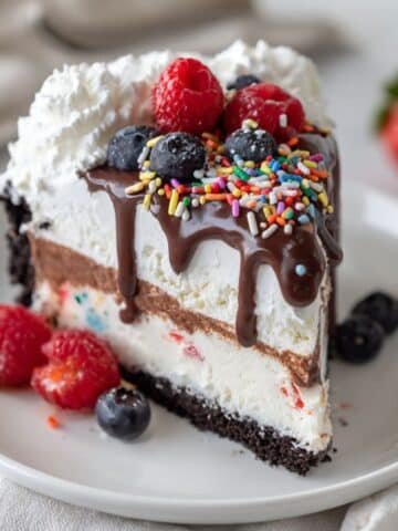 Homemade gluten free ice cream cake with cookie crust, layered ice cream, chocolate ganache, whipped cream, and berries on a white kitchen table, top-down view.