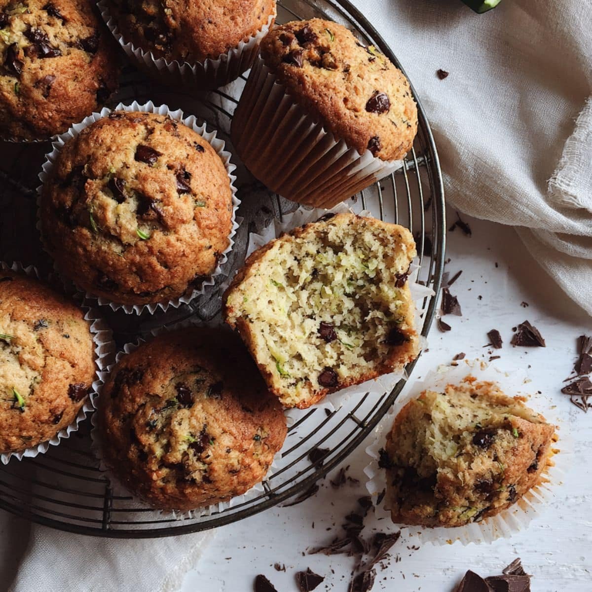 Top-down photo of Zucchini Muffin Chocolate Chip on a white table, one torn open, crumbs and a few chips around.