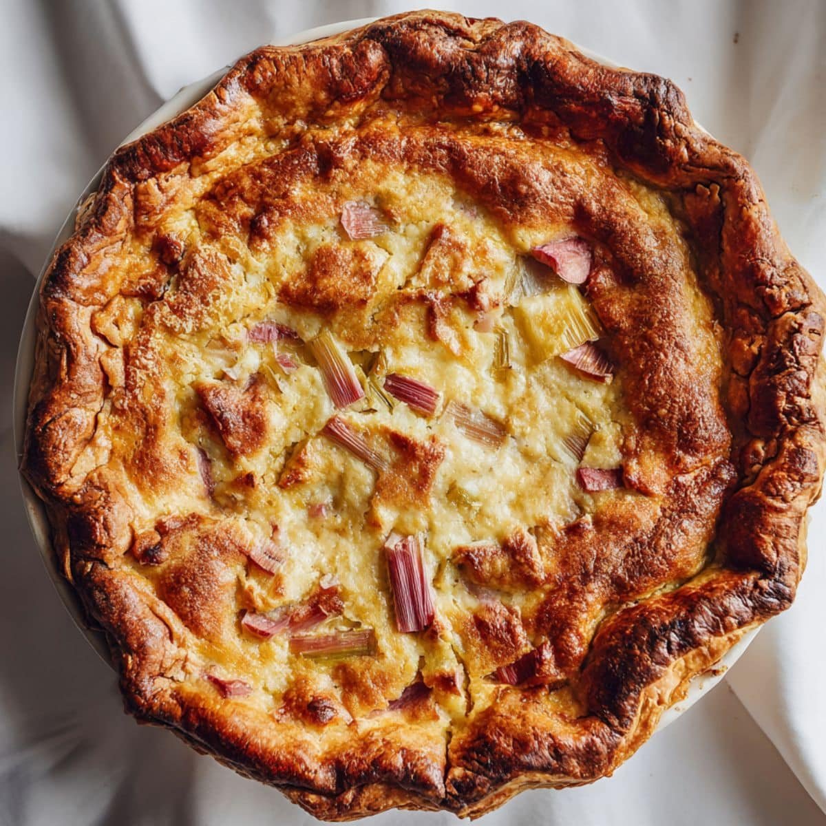 Homemade rhubarb custard pie on a white kitchen table, rustic crust with creamy custard and rhubarb filling, top-down shot.