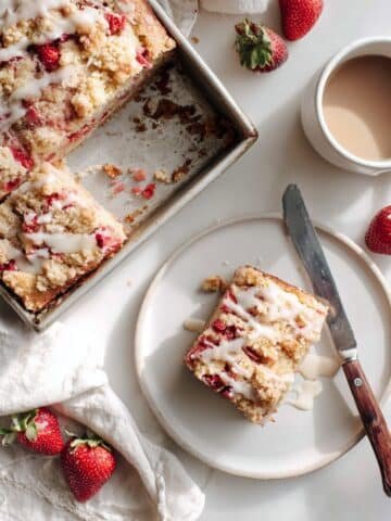Top-down homemade strawberry rhubarb cake in a 9×13 pan on a white table; slice removed, light vanilla glaze, crumbs, natural light.