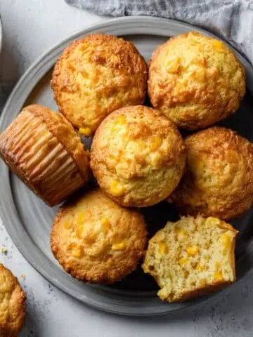 Top-down shot of sweet corn muffins recipe in a tin on a white kitchen table; a few out, crumbs, natural light.