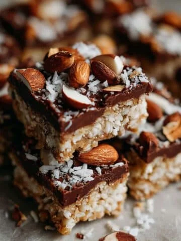 Homemade almond joy bars stacked on a white plate, chocolate coconut almond squares with sea salt on top
