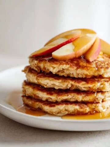 Fluffy homemade apple oat pancakes stacked on a white plate with apple slices and syrup, on a white kitchen counter