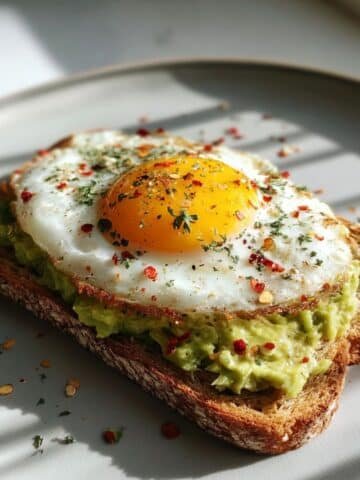 Homemade avocado egg toast on whole grain bread with mashed avocado, fried egg, and herbs on a white kitchen table, photographed from above.