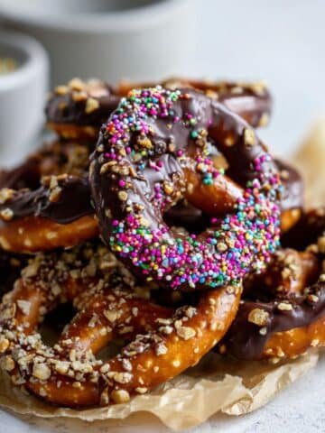 chocolate covered pretzels dipped in milk and dark chocolate with sprinkles and nuts, arranged on parchment paper on a white kitchen table.