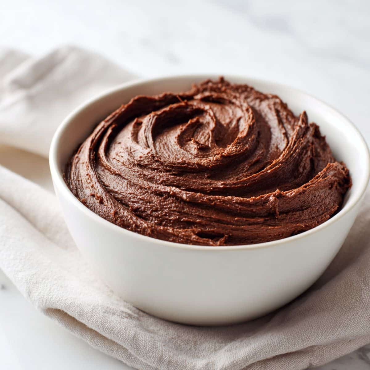 Bowl of creamy homemade chocolate fudge frosting on a white kitchen counter