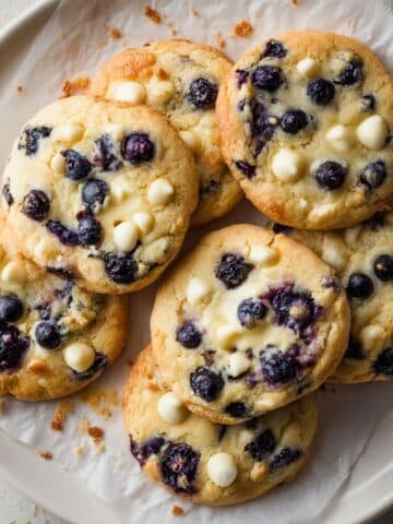 Freshly baked blueberry cheesecake cookies on parchment paper with blueberries and cream cheese filling, shot top-down on a white kitchen table.