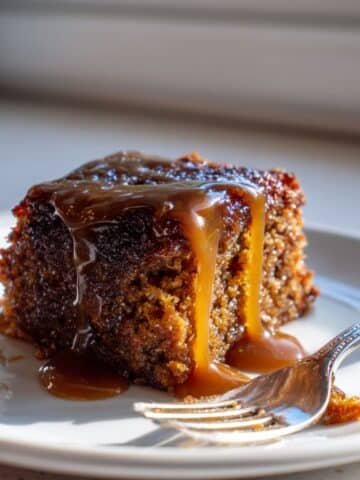 A slice of gluten free sticky toffee pudding drizzled with toffee sauce on a white plate, top-down on a white kitchen table.