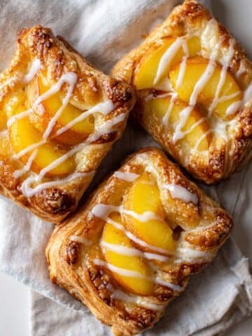 Homemade peach danish pastries on a white kitchen table, golden brown with peach slices and glaze, photographed from above.